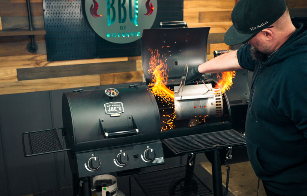 PitMaster loading fire into an Oklahoma Joe's dual function smoker grill combo during an indoor barbecue demonstration